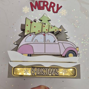 May include: A pink car with a Christmas tree on top, driving through a snowy scene. The car has a gold trim and yellow wheels. The scene is lit up with white lights and the words "Merry Christmas" are displayed on the front of the car.