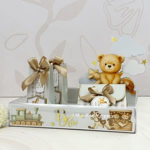 May include: A clear acrylic display case with baby-themed decorations. Features a toy train, teddy bears, and gift bags tied with gold ribbons. The display includes a teddy bear with an airplane and clouds.