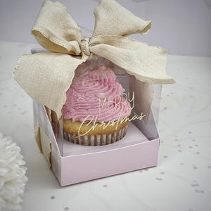 Puede incluir: Un cupcake con glaseado rosa en una caja transparente con una base a cuadros rosa y blanca. La caja está atada con un gran lazo beige. Las palabras "MERRY Christmas" están escritas en la parte delantera de la caja.