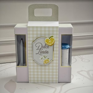 May include: A rectangular light pink gift box with a handle and a central panel featuring a beige gingham pattern. The label reads Dulce Limón and displays lemon slices and white blossoms. Two side windows reveal a grey pen and a white polka dot tube.