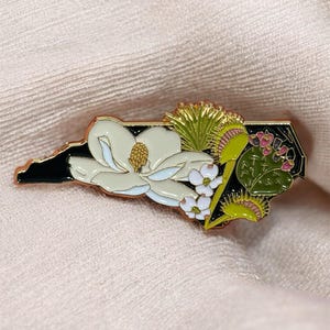 May include: Enamel pin in the shape of North Carolina, featuring a white magnolia flower, small white flowers, and a Venus flytrap. The pin has a black border with gold accents and is set against a peach-colored fabric.