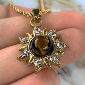 May include: A gold necklace with a sun pendant featuring a tiger's eye stone surrounded by clear crystals.