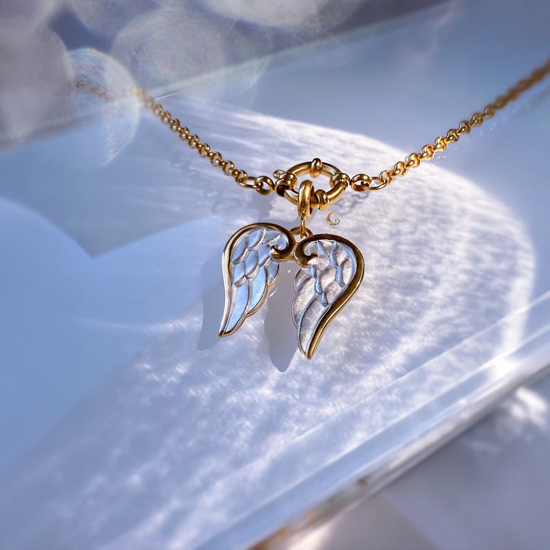 Angel wings necklace with gold chain photographed on marble-like surface in warm sunlight reflections.
