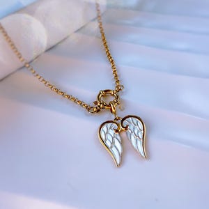 Close-up of gold angel wings pendant necklace placed on stone-like white surface, glowing in soft daylight.