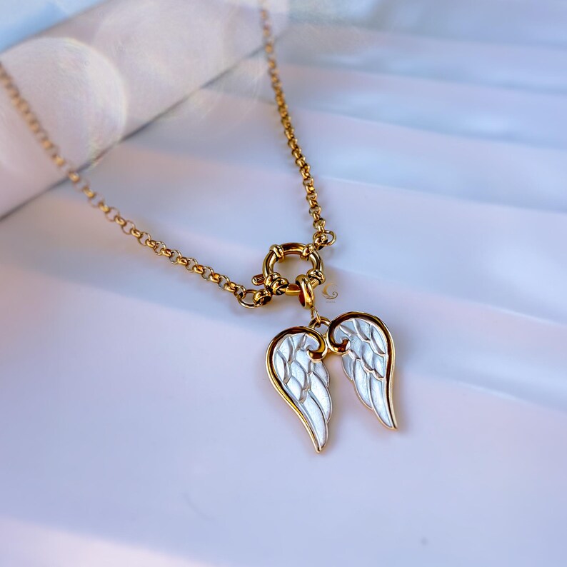 Close-up of gold angel wings pendant necklace placed on stone-like white surface, glowing in soft daylight.