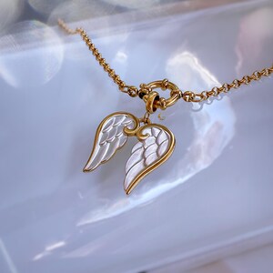 Gold and white angel wings pendant necklace captured on bright surface with soft shadows and reflections.