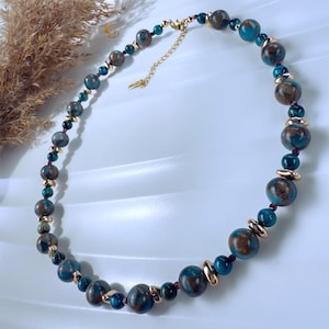 Puede incluir: Collar de cuentas con grandes cuentas redondas en tonos turquesa y marrón, intercaladas con cuentas más pequeñas en tonos turquesa y dorado. El collar tiene una cadena y un cierre dorados. El collar se muestra sobre una superficie blanca.