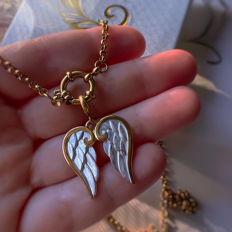 Hand holding gold angel wings necklace, showing double wing pendant with silver enamel and textured details.