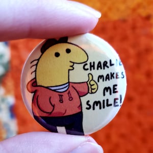 Charlie button against an orange quilt backdrop. Two fingers hold up the small button. The text on the button says Charlie Makes Me Smile!