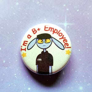 K&ouml;nnte beinhalten: Ein runder Button mit dem Text "I'm a B+ Employee!" in Rot. Der Button zeigt einen Cartoon-Alien-Charakter mit blauer Haut, gelben Augen und einer schwarzen Uniform. Zwei gelbe Sterne sind ebenfalls abgebildet.