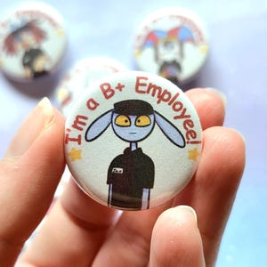 K&ouml;nnte beinhalten: Ein runder Button mit einer Cartoonfigur mit blauer Haut und gelben Augen, die eine schwarze Uniform tr&auml;gt. Der Text auf dem Button lautet "I'm a B+ Employee!". Weitere Buttons mit &auml;hnlichen Designs sind im Hintergrund zu sehen.