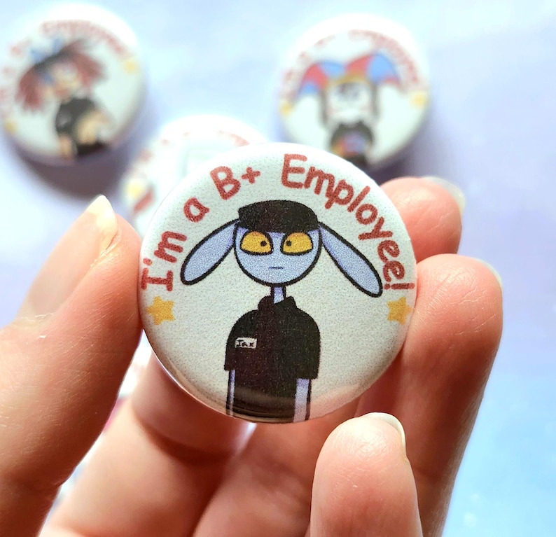 K&ouml;nnte beinhalten: Ein runder Button mit einer Cartoonfigur mit blauer Haut und gelben Augen, die eine schwarze Uniform tr&auml;gt. Der Text auf dem Button lautet "I'm a B+ Employee!". Weitere Buttons mit &auml;hnlichen Designs sind im Hintergrund zu sehen.