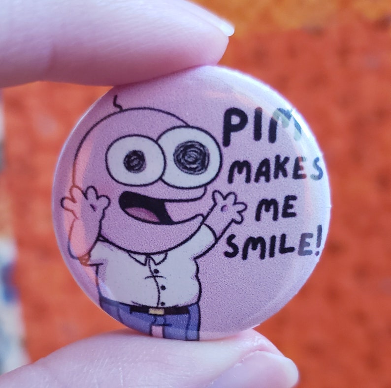 Pim button against an orange quilt backdrop. Two fingers hold up the small button. The text on the button says Pim Makes Me Smile!