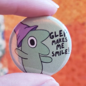 Glep button against an orange quilt backdrop. Two fingers hold up the small button. The text on the button says Glep Makes Me Smile!