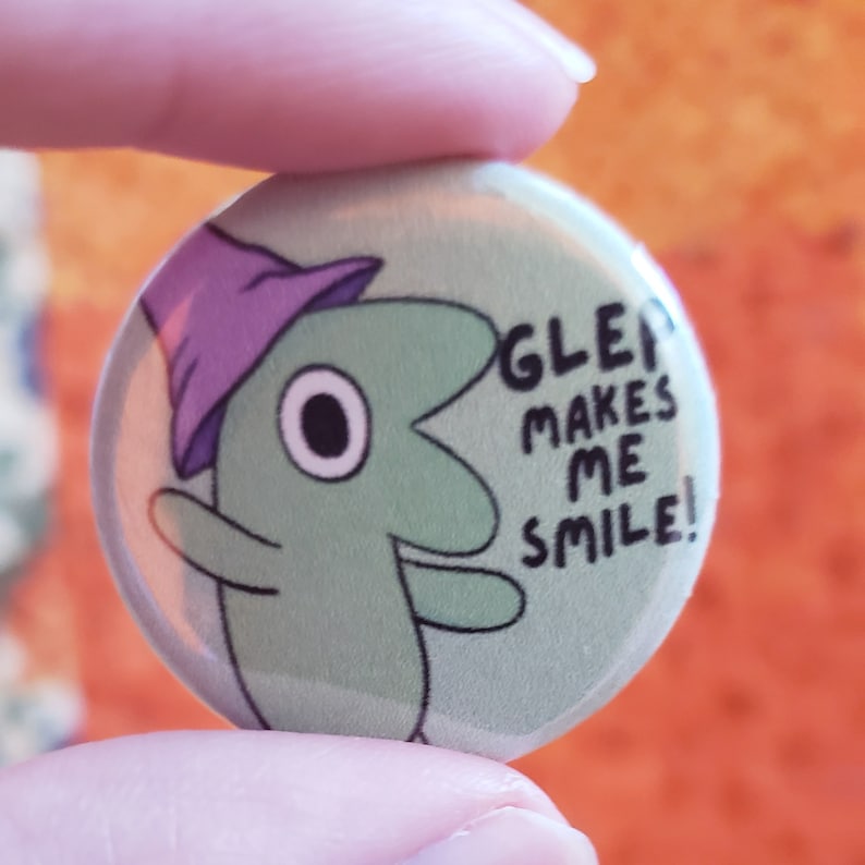 Glep button against an orange quilt backdrop. Two fingers hold up the small button. The text on the button says Glep Makes Me Smile!