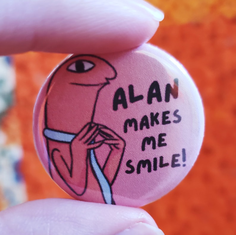 Alan button against an orange quilt backdrop. Two fingers hold up the small button. The text on the button says Alan Makes Me Smile!