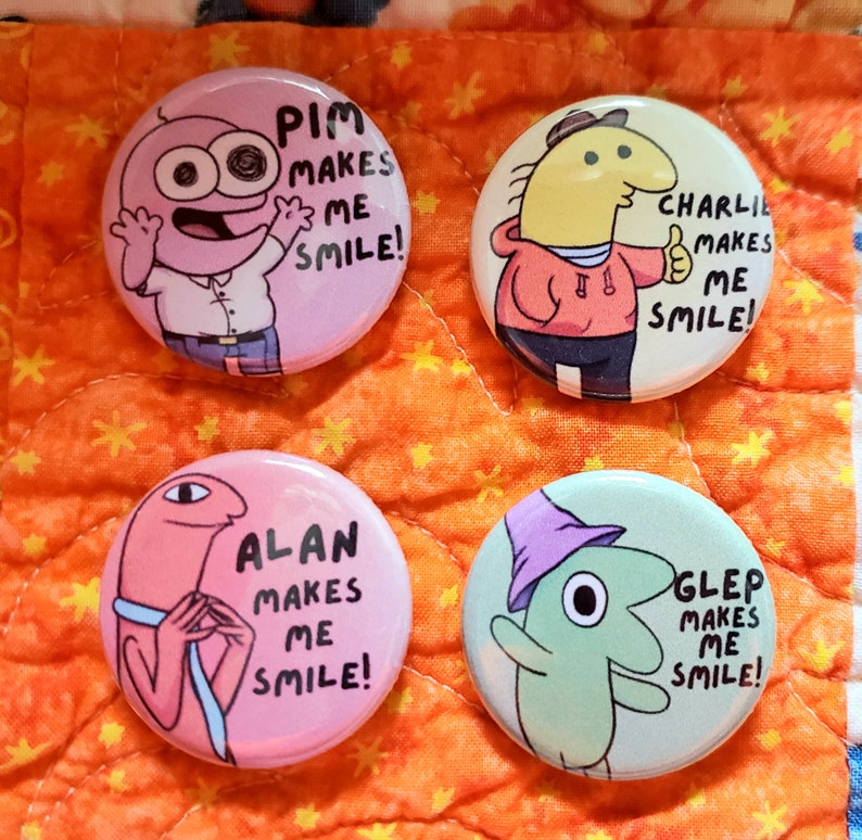 4 Smiling Friends buttons on top of an orange quilt backdrop. Pim and Charlie are on top and Alan and Glep are underneath. Text on the buttons have the charactes names along with Makes Me Smile!