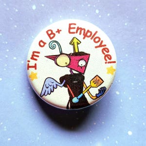 K&ouml;nnte beinhalten: Ein runder Button mit einem Cartoon-Roboter-Design. Der Roboter ist rosa, schwarz und gelb, h&auml;lt einen Spatel und tr&auml;gt einen Fl&uuml;gel. Der Text "I'm a B+ Employee!" ist am Rand aufgedruckt.