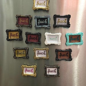 May include: A collection of framed cross-stitch art pieces, each displaying the word "fuck". The frames vary in color, including gold, black, and teal, and are arranged on a metallic surface. The cross-stitch is in various colors.