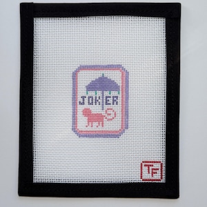 May include: Needlepoint canvas with a black border. The design features the word "JOKER" above a pink lion under a purple umbrella. A small red and white logo is in the bottom right corner.
