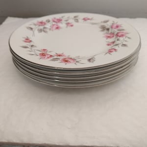 Puede incluir: Una pila de seis platos de porcelana blanca. Cada plato presenta un delicado diseño floral de rosas rosas y hojas grises alrededor del borde. Los platos tienen un borde dorado.