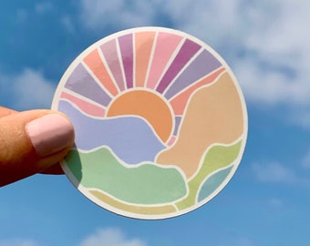 May include: A colorful sticker with a sun rising over a mountain range. The sun is orange, the mountains are purple, blue, green, and pink, and the sky is a light blue.