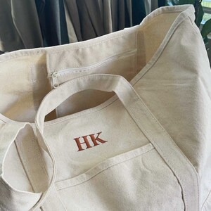 Può includere: Una borsa tote in tela bianca con una tasca con cerniera e due manici. La borsa ha le iniziali "HK" ricamate in arancione sul davanti.