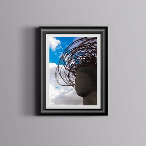 Puede incluir: Una fotografía en blanco y negro de una escultura con una cabeza grande, redonda y gris y una estructura de metal oxidado enredada que se asemeja al cabello. La escultura se coloca contra un cielo azul con nubes blancas.