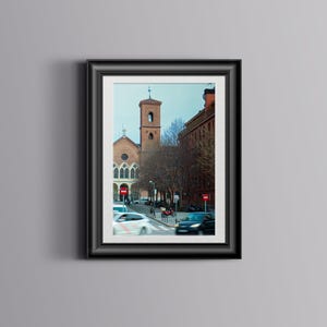 May include: A framed photograph of a European city street scene. The image features a church with a bell tower, red brick buildings, and cars on the street. The sky is a pale blue, and the overall tone is muted.