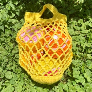 Puede incluir: Una bolsa de malla amarilla tejida a crochet con asa, llena de cuatro bolas tejidas a crochet en tonos naranja, rosa y blanco.