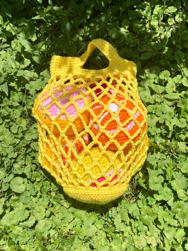 Puede incluir: Una bolsa de malla amarilla tejida a crochet con asa, llena de cuatro bolas tejidas a crochet en tonos naranja, rosa y blanco.