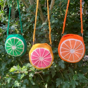 May include: Three crocheted bags with fruit designs. The bags are green, pink, and orange and have a crocheted strap. The bags are hanging from a tree branch.
