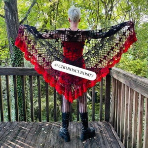 May include: A black and red crochet shawl with a floral design. The shawl has a scalloped edge and is displayed outdoors. The text "CHIAROSCUROSITY" is visible on the shawl.