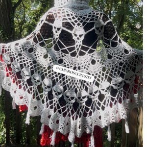 May include: A white crocheted hooded shawl featuring an intricate web pattern and multiple skull motifs integrated into the design. The garment has a scalloped edge with small tassels hanging from the bottom. The text CHIAROSCUROSITY is visible.