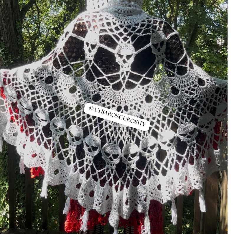 May include: A white crocheted hooded shawl featuring an intricate web pattern and multiple skull motifs integrated into the design. The garment has a scalloped edge with small tassels hanging from the bottom. The text CHIAROSCUROSITY is visible.