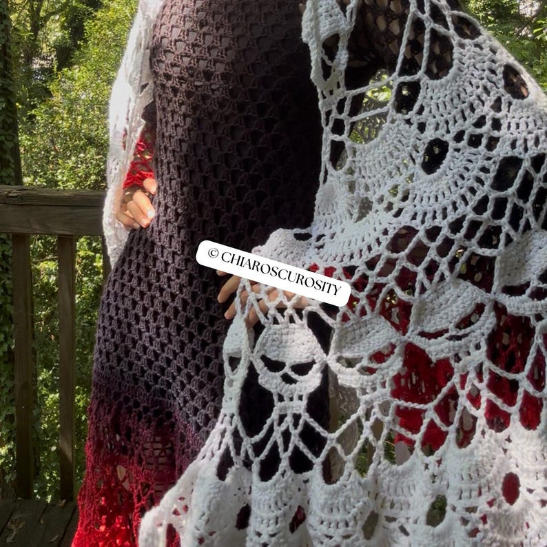 May include: A dark brown and red gradient crochet dress featuring a long-sleeved design with a grid-like pattern. A white crochet shawl with a skull motif is draped over the dress. A white rectangular label displays the text CHIAROSCUROSITY.