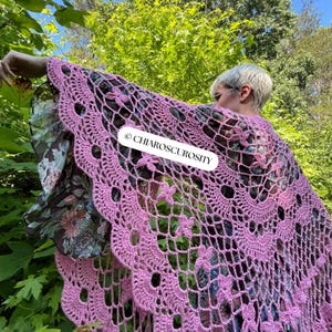 May include: A light purple, open-weave crochet shawl with a floral pattern. The shawl has a scalloped edge and is draped over a floral patterned garment. The text "CHIAROSCUROSITY" is visible on the shawl.