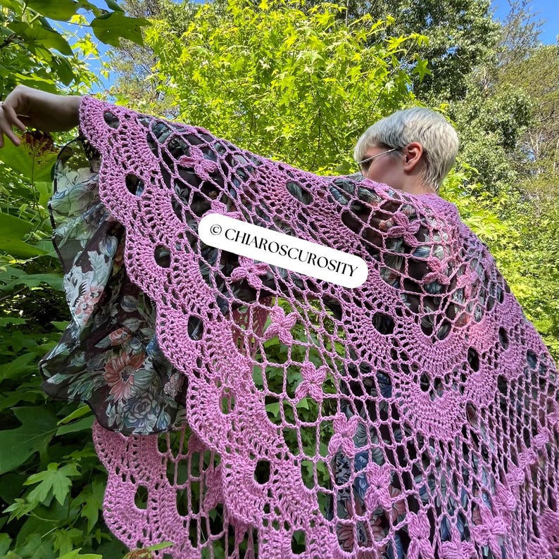 May include: A light purple, open-weave crochet shawl with a floral pattern. The shawl has a scalloped edge and is draped over a floral patterned garment. The text "CHIAROSCUROSITY" is visible on the shawl.