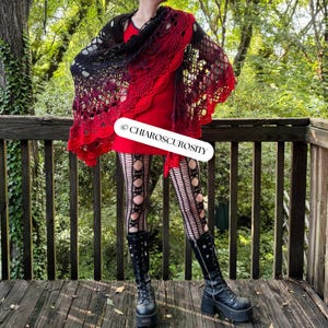 May include: A black to red gradient crocheted shawl worn over a red dress. The image also shows black tights with circular cutouts and black platform boots. The text "&copy; CHIAROSCUROSITY" is visible. The scene is set outdoors.