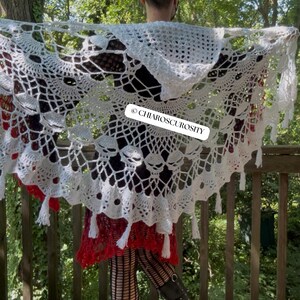 May include: A white crocheted hooded shawl featuring a repeating skull pattern throughout the lace design, finished with a scalloped edge and hanging tassels. A rectangular white label with the black text CHIAROSCUROSITY is attached.