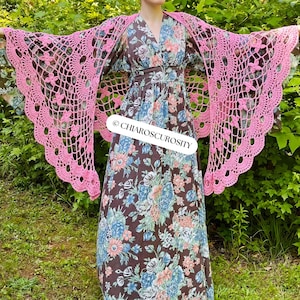 May include: A pink, open-weave crochet shawl with a floral pattern is draped over a long, floral-print dress. The dress is brown with blue and white flowers. The text "CHIAROSCUROSITY" is visible on the dress.