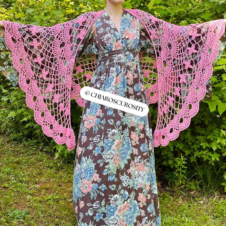 May include: A pink, open-weave crochet shawl with a floral pattern is draped over a long, floral-print dress. The dress is brown with blue and white flowers. The text "CHIAROSCUROSITY" is visible on the dress.