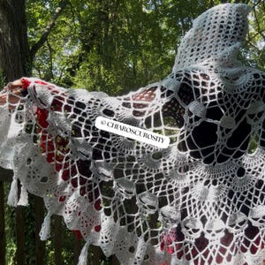May include: A white crocheted hooded shawl featuring an intricate web-like pattern with skull motifs and a scalloped edge with tassels. A red layer is visible underneath the white crochet. The text CHIAROSCUROSITY is printed on a white label.