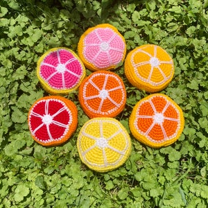 Puede incluir: Siete rodajas de fruta de ganchillo en varios tonos de naranja, amarillo y rosa. Las rodajas est&aacute;n dispuestas en c&iacute;rculo sobre un lecho de hierba verde.
