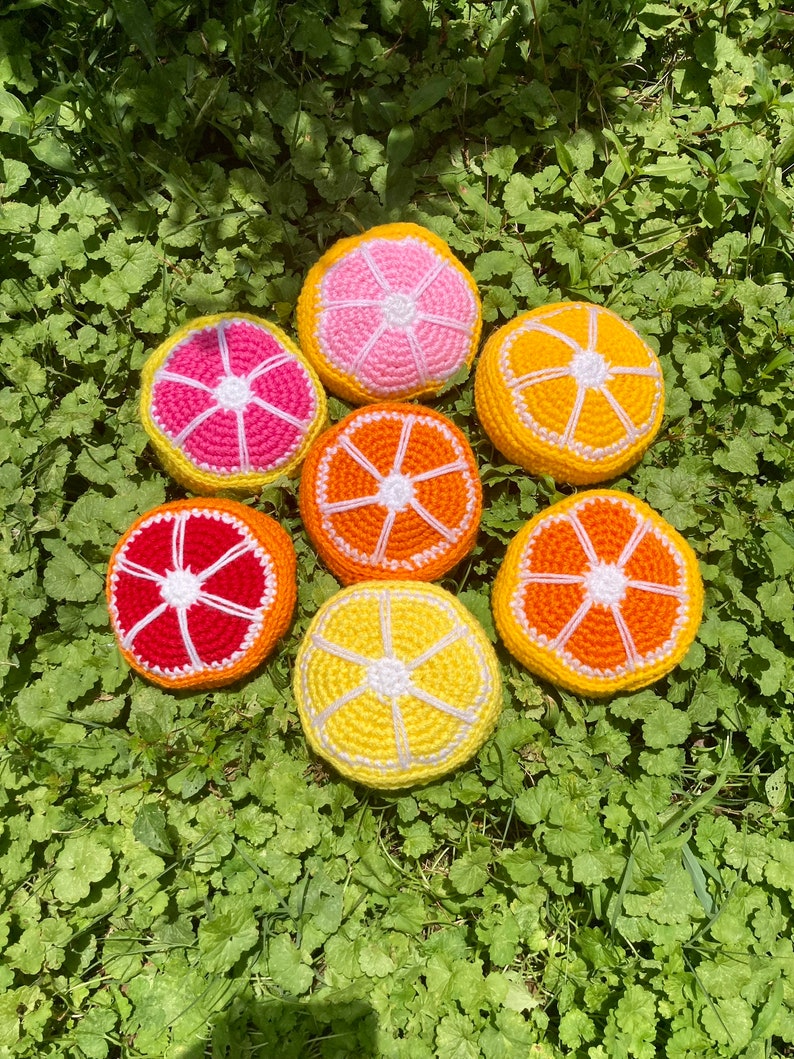 Puede incluir: Siete rodajas de fruta de ganchillo en varios tonos de naranja, amarillo y rosa. Las rodajas est&aacute;n dispuestas en c&iacute;rculo sobre un lecho de hierba verde.