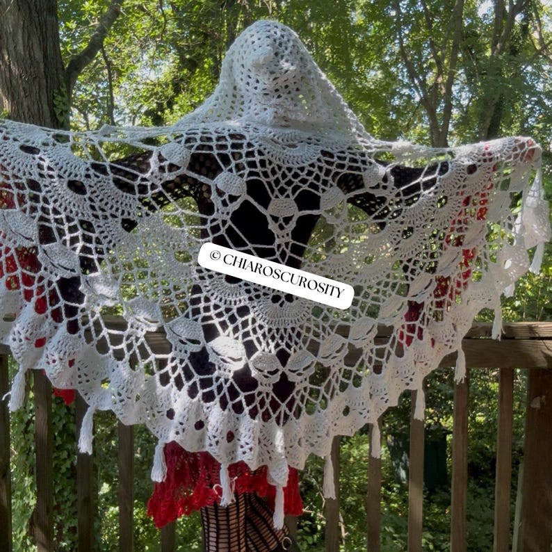 May include: A white hooded crochet shawl featuring an intricate web-like pattern with skull motifs throughout the design. The garment includes a scalloped edge with small tassels and a rectangular label with the text CHIAROSCUROSITY.