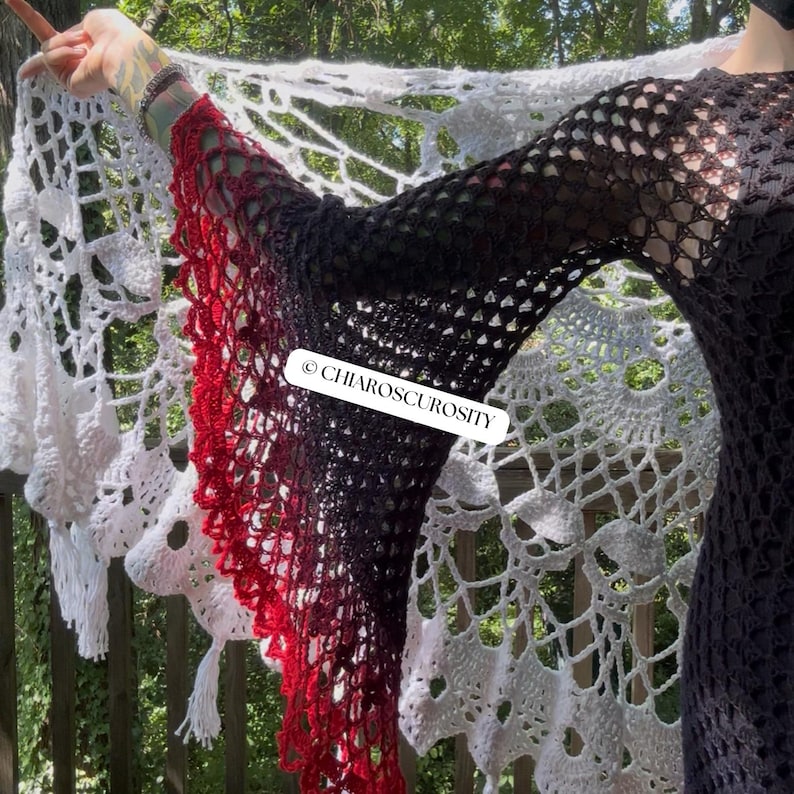 May include: A long-sleeved crochet top featuring a black bodice and sleeves that transition into a vibrant red gradient at the flared cuffs. A white, intricate, large-scale crochet shawl with a skull motif is draped behind. Text reads CHIAROSCUROSITY.