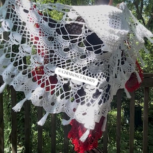 May include: A white crocheted shawl featuring an intricate web-like pattern with skull motifs and small tassels along the scalloped edge. The fabric displays the text CHIAROSCUROSITY in black letters. The design is layered over a red crocheted garment.