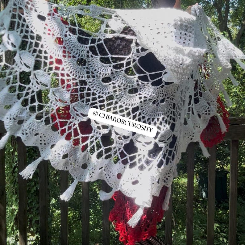 May include: A white crocheted shawl featuring an intricate web-like pattern with skull motifs and small tassels along the scalloped edge. The fabric displays the text CHIAROSCUROSITY in black letters. The design is layered over a red crocheted garment.