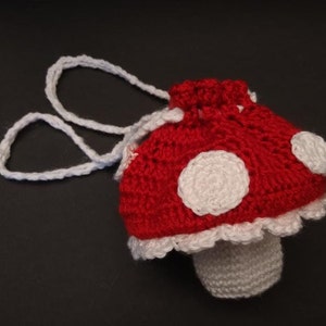 May include: A crocheted red and white mushroom-shaped bag with a white drawstring and white polka dots. The bag has a white crocheted stem.